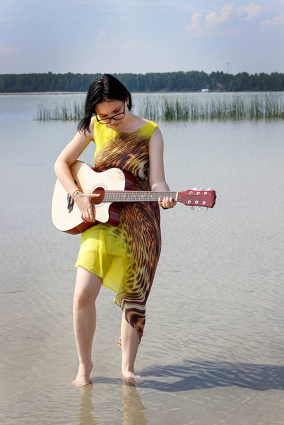 A girl in a dress plays the guitar standing barefoot in the water of a lake in nature. The vertical frame. - Photo, Image