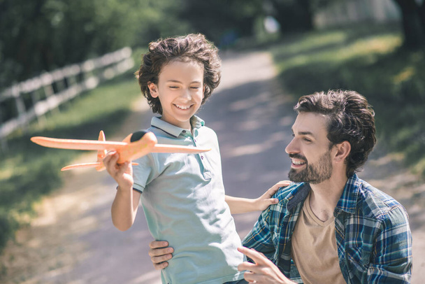 Happy dark-haired boy holding a toy plane and touching his fathers shoulder - Foto, Imagen