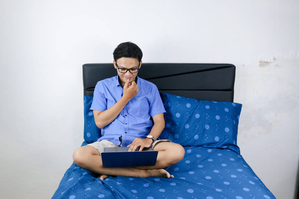 Young asian man is smile and enjoy when working at home. A man sitting on bed and working with laptop computer. Work from home concept. - Fotoğraf, Görsel