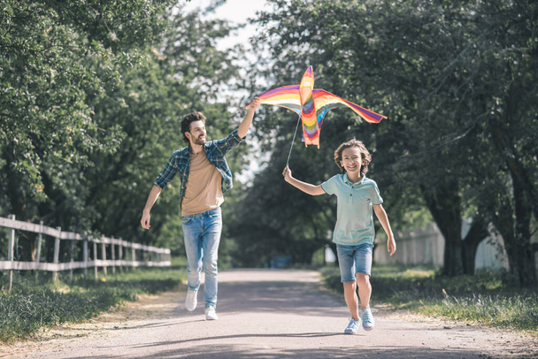 Dark-haired boy and his father running after the colorful kite and looking excited - Fotoğraf, Görsel