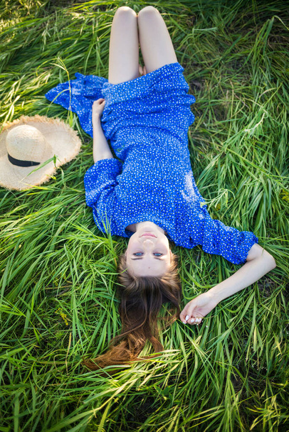 young european girl in a blue dress lies in the grass with a hat - 写真・画像
