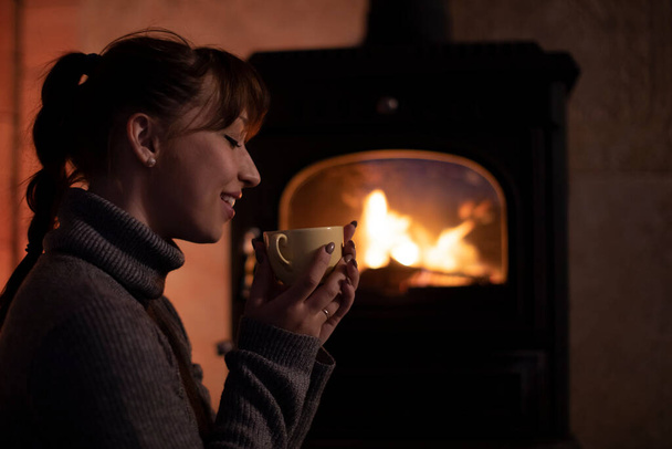 Portrait of young girl in warm sweater drinking hot drink from mug by fireplace - Photo, Image