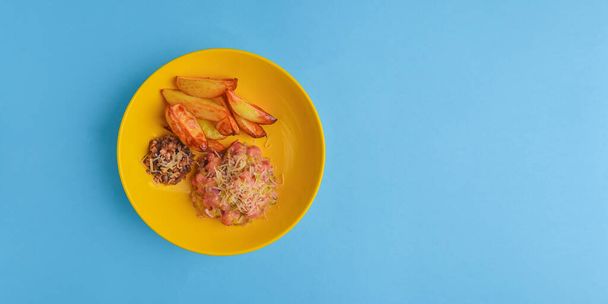 Baked potato with ham and grated cheeses served in a yellow bowl over pastel blue background. - Foto, imagen