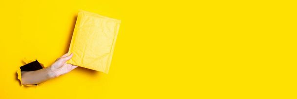 Γυναικείο χέρι κρατά ένα κίτρινο φάκελο ταχυδρομείου σε σκισμένο κίτρινο φόντο. Μπάνερ - Φωτογραφία, εικόνα