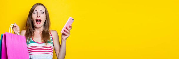 Surprised shocked young girl with phone holding shopping bags on yellow background. Banner - Photo, Image
