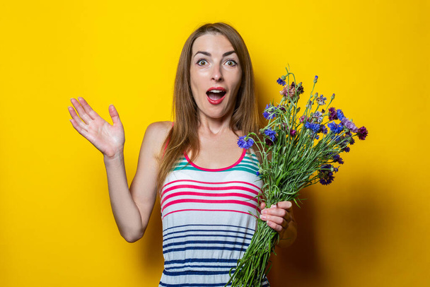 Удивлённая шокированная молодая женщина в полосатом платье с полевыми цветами на жёлтом фоне - Фото, изображение