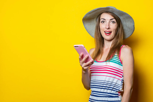Happy young woman holding phone, wearing hat and striped dress posing on yellow background - Fotó, kép