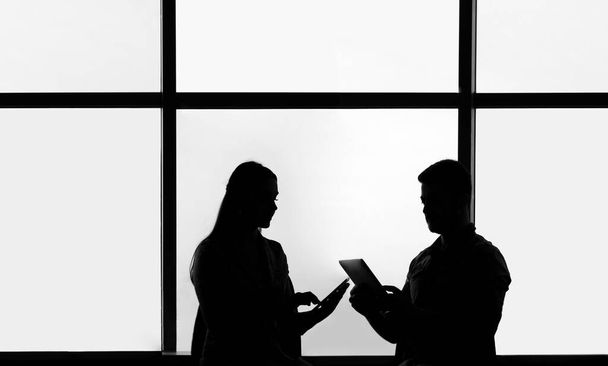 Silhouettes of 2 two business people man and woman in office interior, man showing information on screen of his tablet to his colleague - Foto, Bild