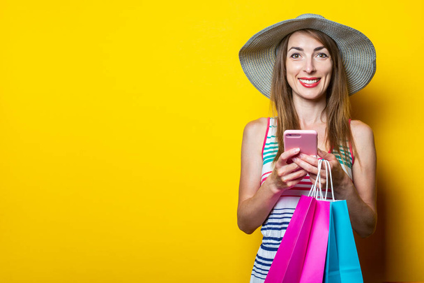 Šťastná mladá dívka s telefonem drží balíčky s nákupy na žlutém pozadí - Fotografie, Obrázek
