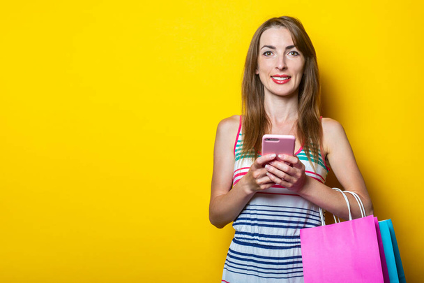 Krásná mladá žena drží telefon v ruce, s balíčky s nákupy na žlutém pozadí - Fotografie, Obrázek