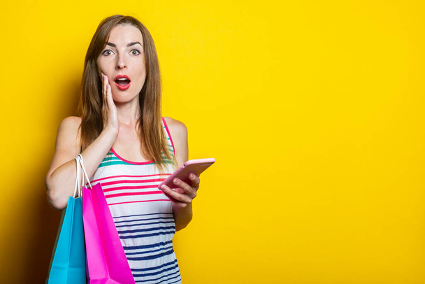 Shocked young woman holding palms on her cheek, with phone and bags of purchases on yellow background - Foto, imagen