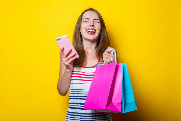 Joyful young girl with phone holding shopping bags on yellow background - Фото, изображение
