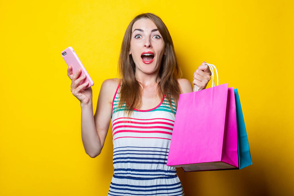 Έκπληκτη σοκαρισμένη νεαρή γυναίκα με τηλέφωνο κρατώντας τσάντες για ψώνια σε κίτρινο φόντο - Φωτογραφία, εικόνα
