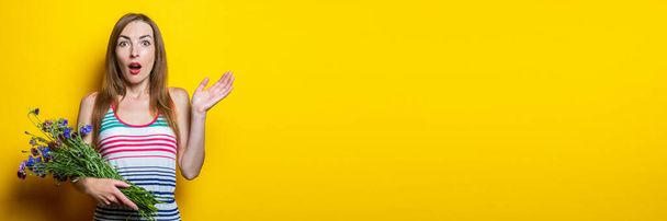 Surprised shocked young woman in striped dress holding wildflowers on yellow background. Banner - 写真・画像