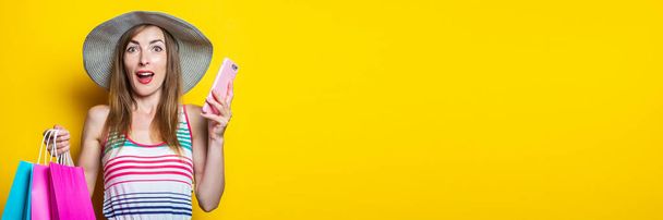 Ξαφνιάστηκα που μια νεαρή γυναίκα με καπέλο κρατούσε τηλέφωνο και τσάντες για ψώνια σε κίτρινο φόντο. Μπάνερ - Φωτογραφία, εικόνα