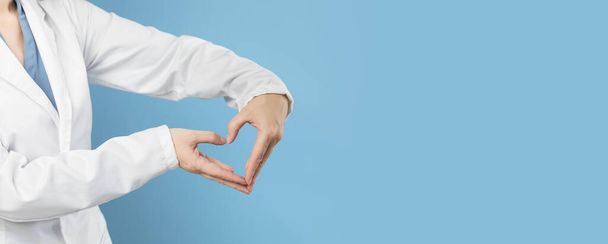 Female doctor in white uniform forms a heart shape with her hands. Minimal blue background. Banner with copy space. Heart, cardiology and medical care support and assistance and love - Фото, изображение