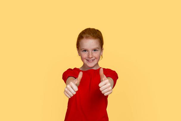 Waist up emotional portrait of a young smiling girl with red hair wearing red shirt  showing approving gesture with hands against  yellow background in the studio with copy space.  - Photo, Image
