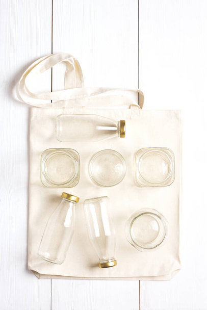 Afvalvrije winkelset. Een herbruikbare geweven tas rust op een wit oppervlak met glazen flessen en potten erop. Milieuvriendelijke levensstijl. Verticale oriëntatie, knollen. - Foto, afbeelding