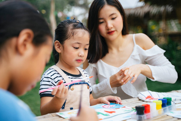 Asiatische große Schwester hilft kleiner Schwester beim Malen von Aquarell auf Papier im Freien. - Foto, Bild