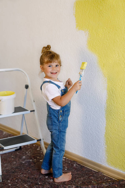 Симпатичная маленькая девочка, красит стену желтым цветом в новом доме. Семейный ремонт многоквартирного дома. Счастливая девочка рисует стены, выбирая цвет палитрой, развлекаясь кистью, в помещении - Фото, изображение