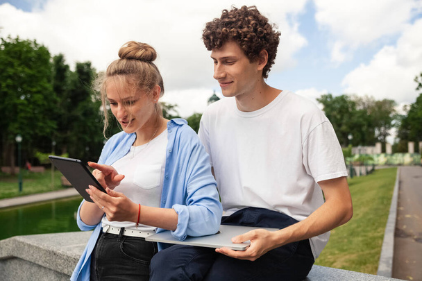 Image of attractive European freelance people man and woman in urban park in smart casual style, using technology for working outdoor together on tablet computer. - Photo, Image