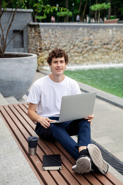 Portrait young European man in wireless earphones wearing white t-shirt sitting in a city park and working on laptop. Freelance work concept outdoors.  - Photo, Image