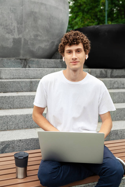 Education, technology and internet concept - European young man in wireless earphones wearing white t-shirt with laptop computer studying outdoor in city park at summer day and listening to music.  - Photo, Image