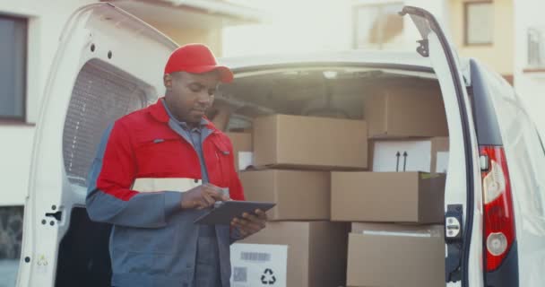 Afroameričtí mladí pošťáci v červeném kostýmu a čepici počítají poštovní schránky v dodávce s tabletovým zařízením v ruce. Venku. - Záběry, video