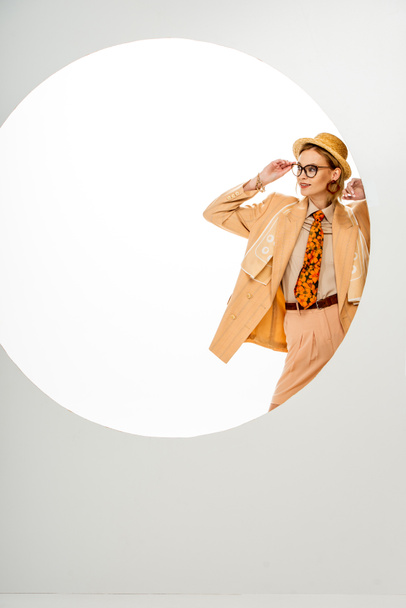 Стильная женщина в соломенной шляпе и бежевой куртке регулирует очки возле круга на белом фоне  - Фото, изображение