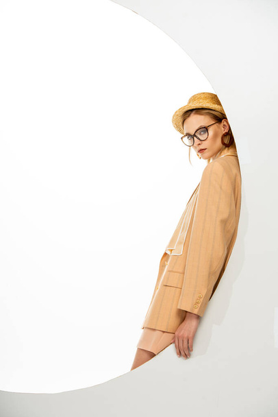 Side view of stylish woman in straw hat and beige jacket posing near circle on white background  - Fotó, kép