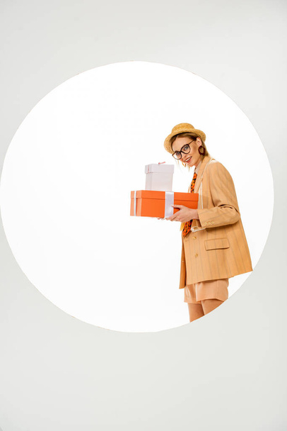 Stylish girl smiling at camera and holding gift boxes behind circle on white background  - Foto, afbeelding
