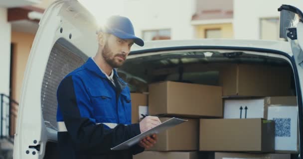 Portrait of the Caucasian good looking male worker of shipping company standing at the van with boxes and writing the document, then smiling to the camera. Outdoor. - Footage, Video