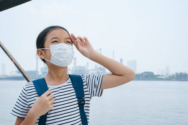 Virus suoja käsite - aasialainen pieni tyttö yllään suojaava naamio, seisoo lähellä joen savusumu päivä. Saasteita joka paikassa. Lakaiseva hiki otsalla. - Valokuva, kuva
