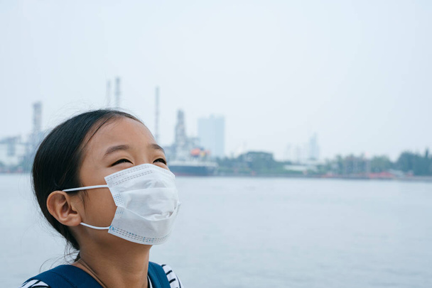 concepto de protección contra el virus Alegre niña asiática con máscara protectora, de pie cerca del río en el día del smog. Contaminación en todas partes. Ella no sabe nada acerca de lo peligroso que es PM2.5
. - Foto, imagen