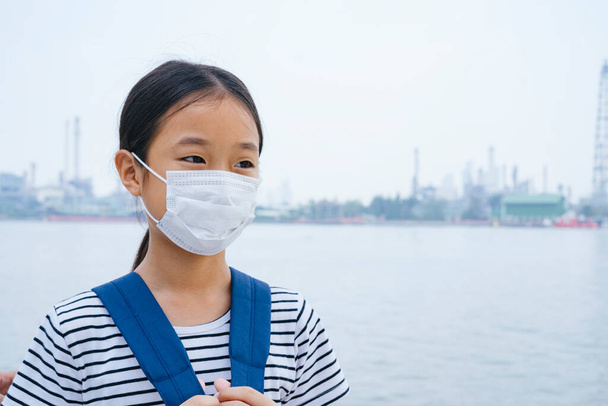 concepto de protección contra el virus - niña asiática con máscara protectora, de pie cerca del río en el día del smog. Contaminación en todas partes
. - Foto, Imagen