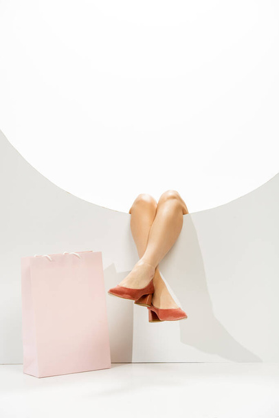 Cropped view of female legs in coral shoes in round hole near shopping bag on white background  - Foto, afbeelding