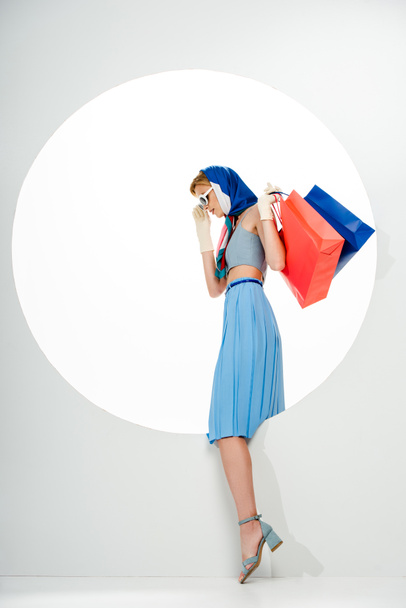Vista lateral de la mujer de moda sosteniendo bolsas de compras rojas y azules cerca de agujero redondo sobre fondo blanco
  - Foto, Imagen