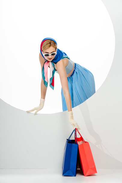 Trendy woman in headscarf and sunglasses putting red and blue shopping bags near circle on white background  - Zdjęcie, obraz