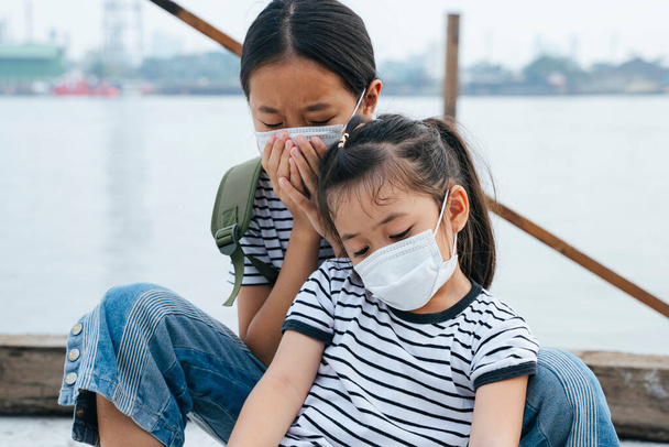 Virus suoja käsite - aasialainen pieni tyttö yllään suojaava naamio, seisoo lähellä joen savusumu päivä. Saasteita joka paikassa. Kaksi siskoa pahoin sairastuu PM 2.5 Polltion. - Valokuva, kuva
