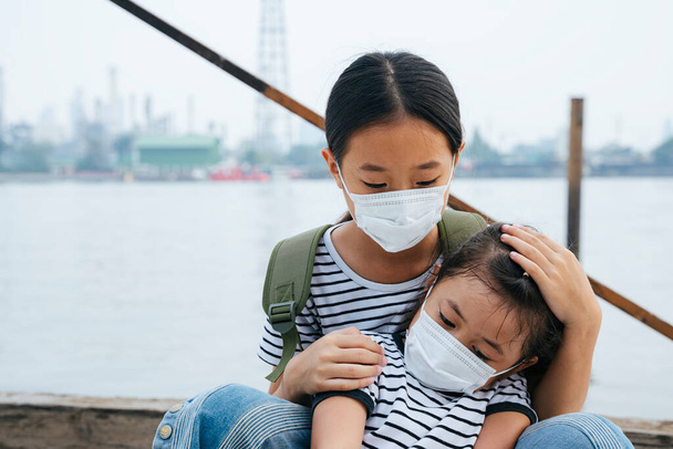 ウイルス保護の概念-アジアの女の子の保護マスクを着て、煙の日に川の近くに立っている。どこでも汚染だ。タイの姉は妹を抱きかかえている。親切さ。家族の愛。人々の世話をする。外科用マスクをした二人の姉妹. - 写真・画像