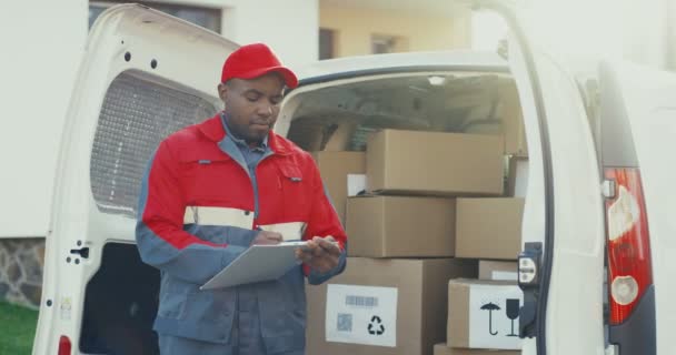 African American mailman in the red uniform and cap standing at the white van with carton boxes and filling in documents on the clipboard. Outdoor. - Footage, Video
