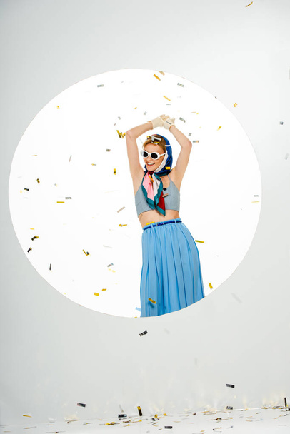Smiling woman in headscarf and sunglasses standing under falling confetti near round hole on white background - Photo, Image