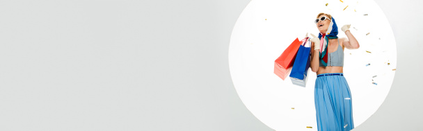 Imagen horizontal de chica con estilo positivo sosteniendo bolsas de compras rojas y azules bajo confeti cayendo cerca del círculo sobre fondo blanco
 - Foto, Imagen
