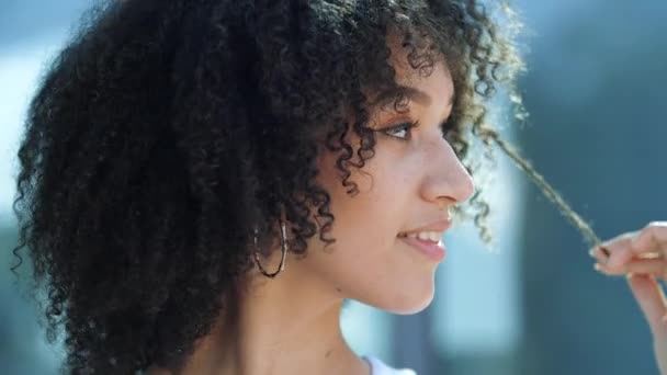 Face portrait of cute positive pretty african young brunette woman model with long curly hairstyle smiling at camera pulling tip of hair with tempting look outdoors in city, slow motion, closeup view. - Footage, Video