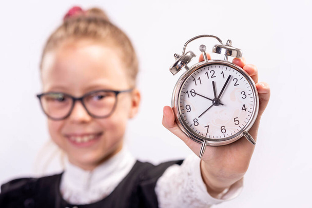 Маленька школярка в окулярах стоїть з годинником на білому тлі. Скоро повернемося до школи
. - Фото, зображення