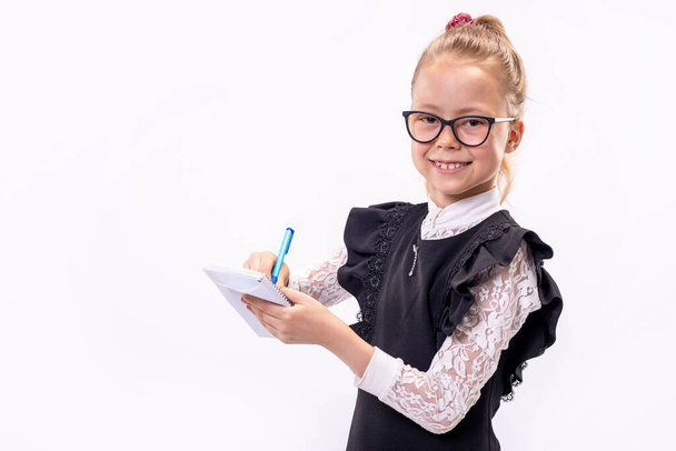 Little schoolgirl in glasses stands on a white background and holds a notebook and pen, copy of the free space. Back to school soon. - Foto, immagini