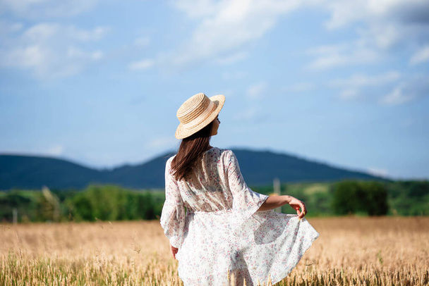 Πορτραίτο ενός κοριτσιού από πίσω σε χωράφι με σιτάρι. Πορτρέτο ενός όμορφου κοριτσιού με λευκό φόρεμα και καπέλο σε χωράφι με σιτάρι. Κορίτσι με λευκό φόρεμα και καπέλο. Πεδίο σίτου. - Φωτογραφία, εικόνα