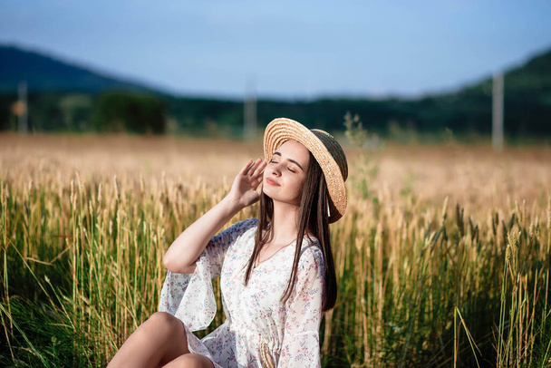 Retrato de una niña en un campo de trigo. La chica está acostada sobre una manta. Retrato de una hermosa niña con un vestido blanco y sombrero en un campo de trigo. Chica con un vestido blanco y sombrero. Campo de trigo
. - Foto, Imagen