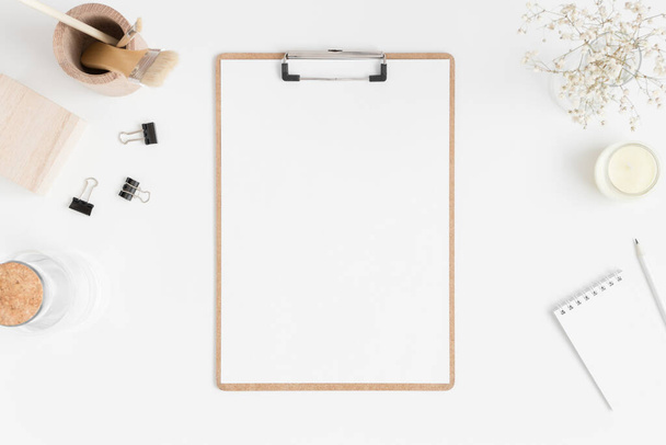 Top view of a wooden clipboard mockup with a gypsophila in vase and workspace accessories on a white table. - Photo, Image