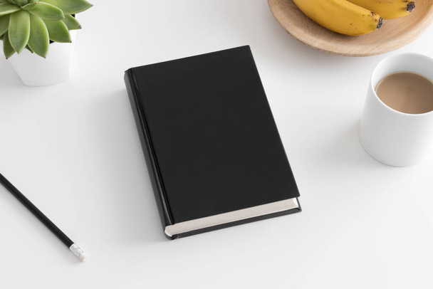 Vista superior de um maquete de livro preto com uma planta suculenta e uma xícara de café em uma mesa branca
. - Foto, Imagem